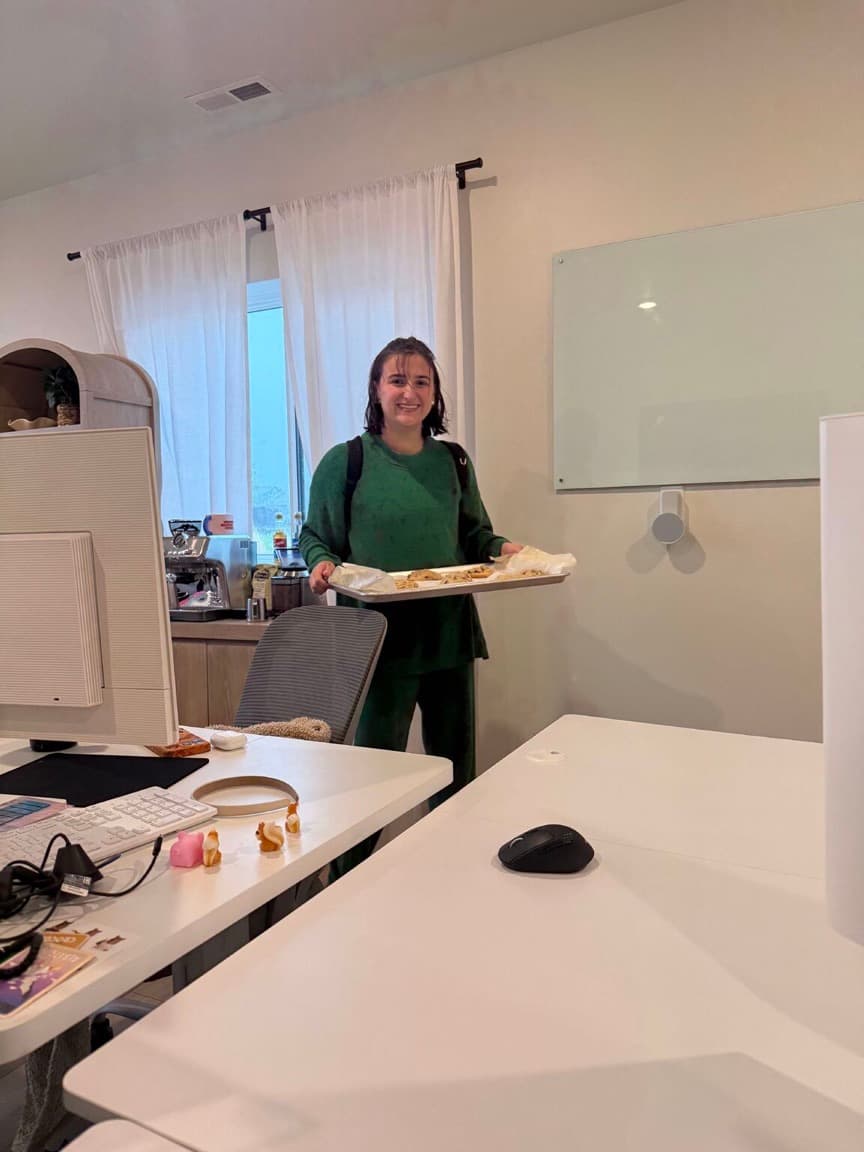 Sam, in a green sweater with her hair still wet from the rain, smiles as she carries a baking sheet of warm cookies into the office.